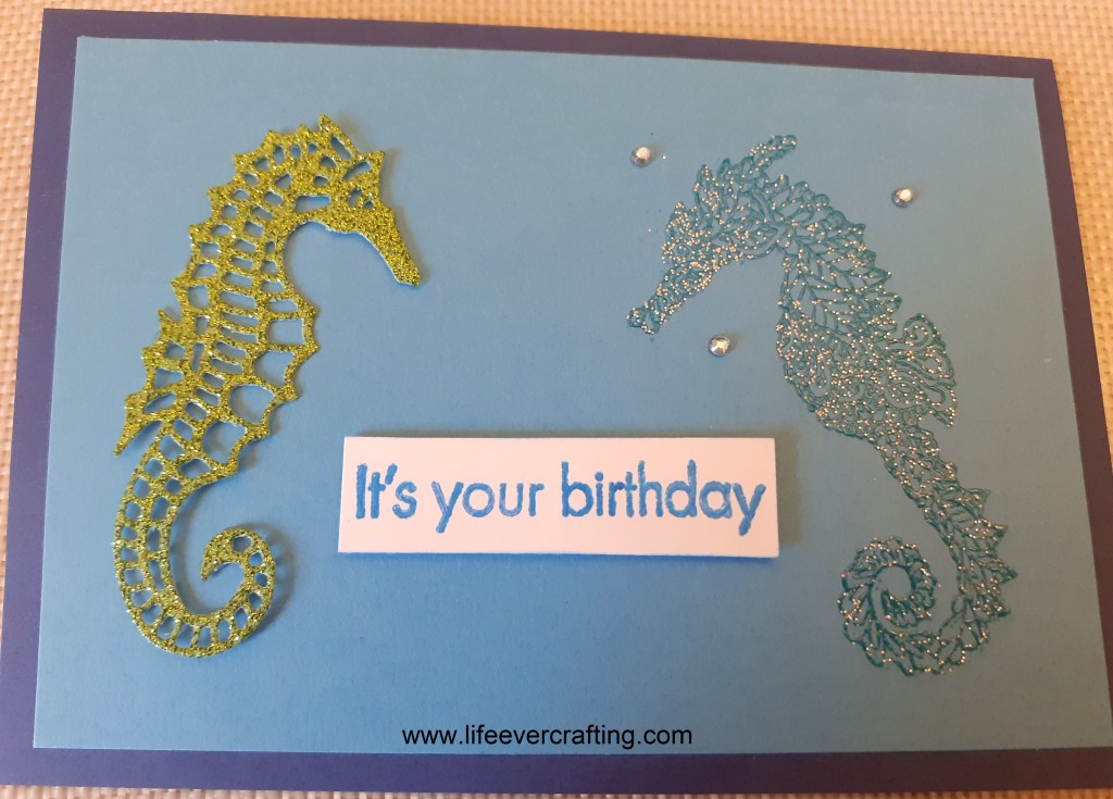 The image is of a blue card with two seahorses facing each other. The one on the left is cut from green glitter card and the one on the right is a glittery dark blue. There are three blue gem dotted around the head of the one on the right and there is a sentiment between them reading It's your birthday.
