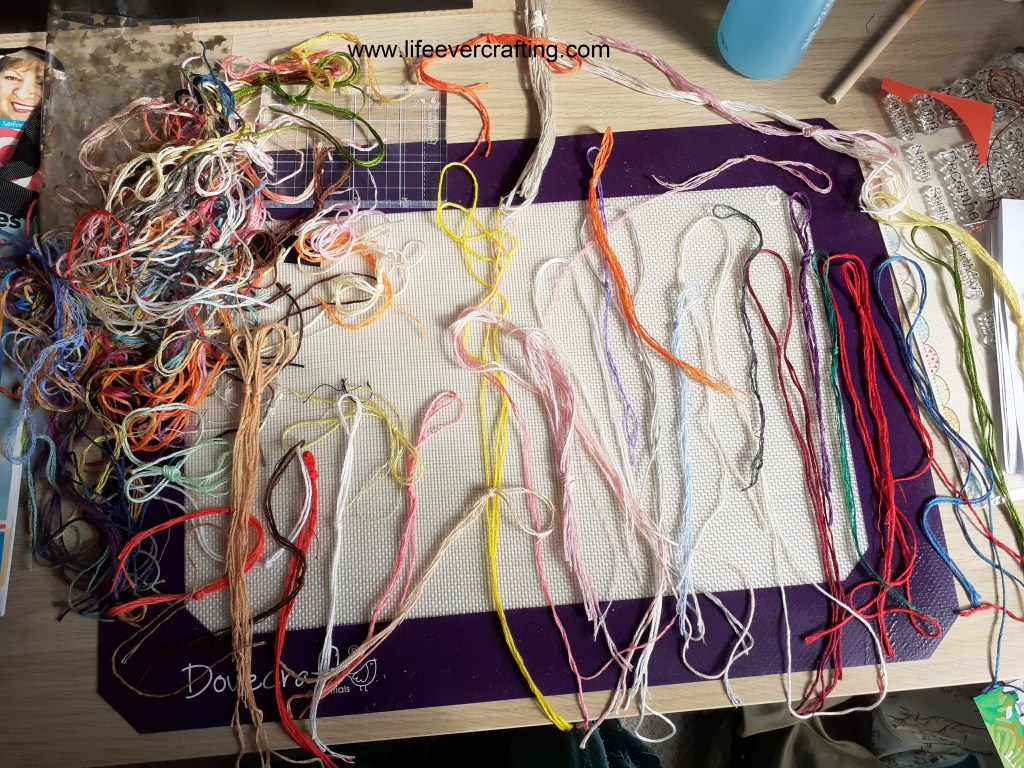 The image shows a desk with skeins of different coloured embroidery threads laid out and a pile of threads in the top left corner.
