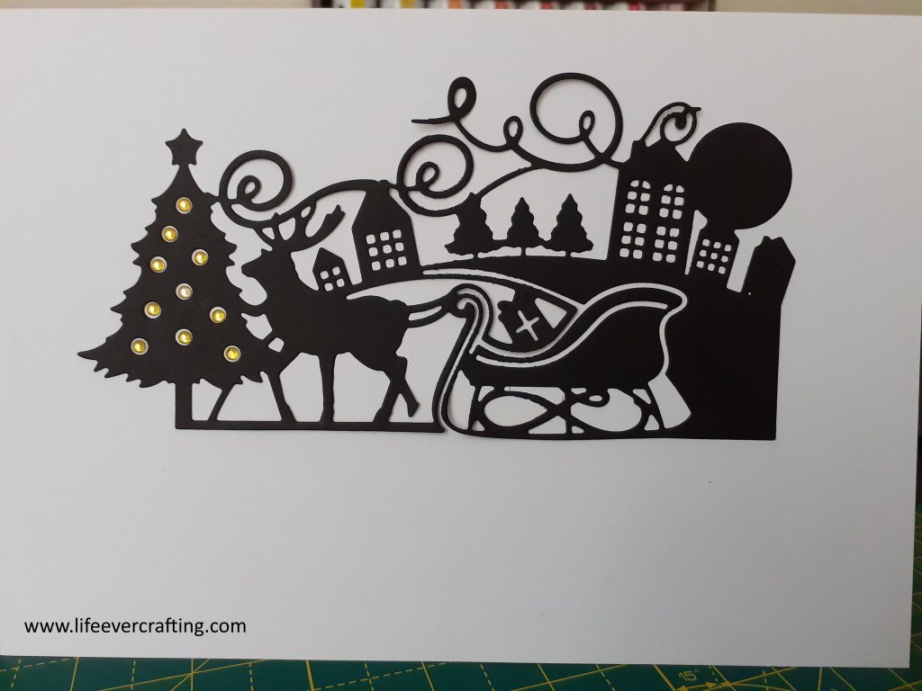 Image shows a Christmas scene with a sleigh drawn by a reindeer standing by a Christmas tree, with houses in the background.