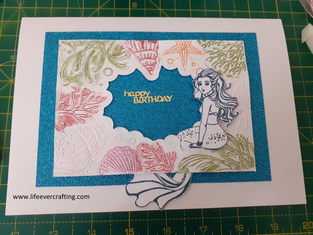 The image shows a birthday card with a glittery blue background. There is a frame of seashells and seaweed in the centre, with a mermaid sitting in the bottom right corner.