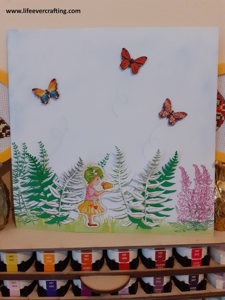 Image shows a fairy holding a snail and standing amongst ferns and foxgloves. Three butterflies are flying above the ferns,