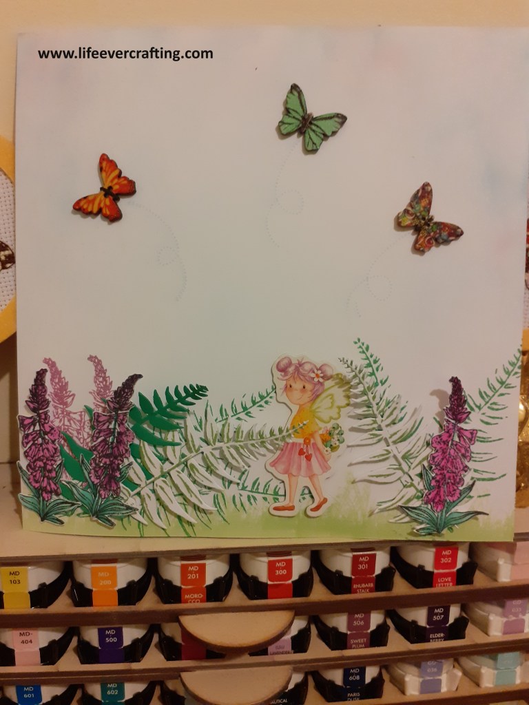 Image shows a fairy walking through ferns and foxgloves, holding a bunch of flowers. Three butterflies are flying in the sky.