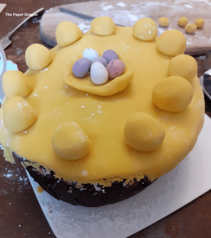 Image shows a fruit cake covered in marzipan, with marzipan balls around the edge and a marzipan nest with mini eggs in the centre.